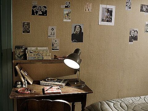 Ein Schreibtisch vor einer tapezierten Wand mit angeklebten Bildern, daneben ein Bett.