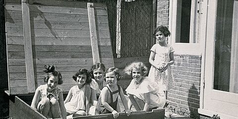 Sechs Kinder in Sommerkleidern in einem Sandkasten, dahinter ein Haus.
