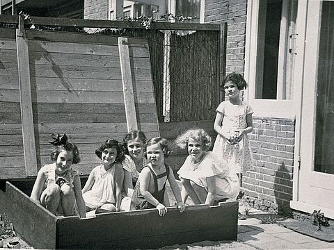 Sechs Kinder in Sommerkleidern in einem Sandkasten, dahinter ein Haus.