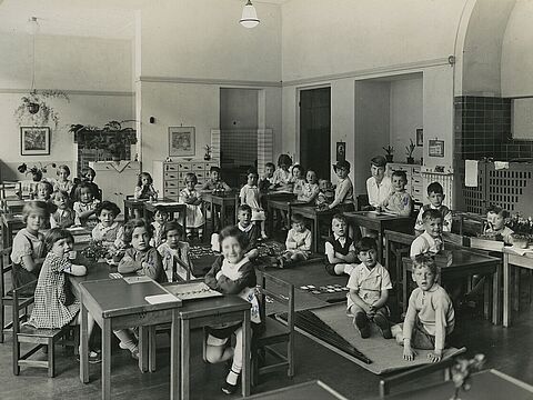 Rund 30 Kinder sitzen an Tischen und auf dem Boden in einem großen Klassenzimmer.