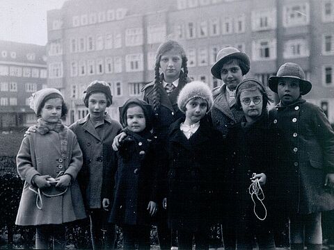 Gruppenbild von acht Kindern in Winterkleidung, dahinter hohe Wohnhäuser.
