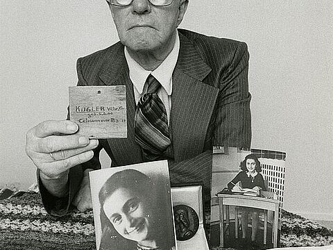 Ein älterer Mann mit Brille an einem Tisch hält ein Holzschild, vor ihm Fotos und Bücher über Anne Frank.