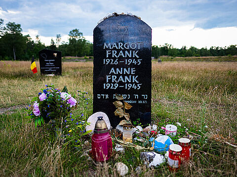 Ein schwarzer Grabstein mit weißer Inschrift auf einer Wiese, davor bunte Blumen und Kerzen.