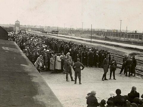Hunderte Menschen stehen in zwei Reihen neben Gleisen, davor uniformierte Personen.