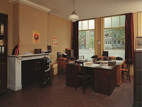 Ein Raum mit drei alten Schreibtischen aus Holz, einem Kamin und großem Fenster.
