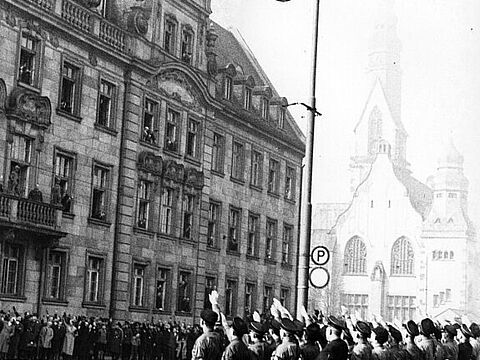 Gruppe von Uniformierten zeigt Hitlergruß zum Balkon eines prächtigen Gebäudes.